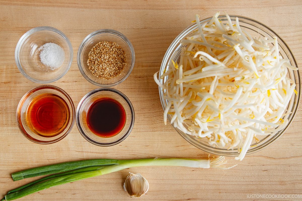 A bowl of bean sprouts sits on a wooden surface next to a green onion, garlic, soy sauce, sesame oil, sesame seeds, and salt in small glass bowls, all neatly arranged for preparing a fresh Bean Sprout Salad.