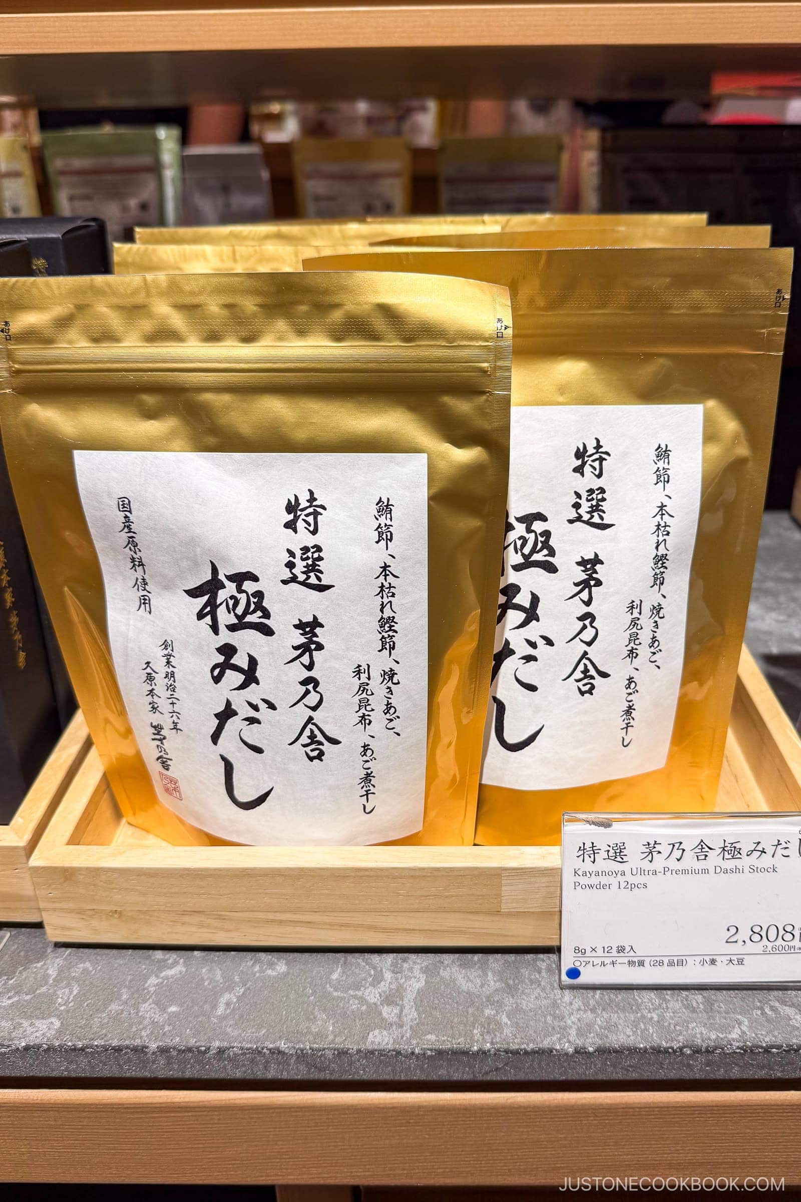 Two gold and white pouches of Kayanoya Ultra-Premium Dashi Stock, are displayed upright in a wooden tray, with a small product label in Japanese and English in front.