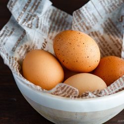 Four brown eggs rest in a bowl lined with printed newspaper, set on a dark wooden surface—a perfect setting to learn how to pasteurize eggs at home before using them in your favorite recipes.