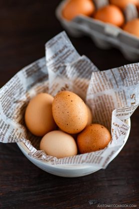 Four brown eggs sit in a white bowl lined with newspaper-print paper. In the background, more eggs are visible in a carton on a dark wooden surface, evoking the careful selection needed for recipes that may require learning how to pasteurize eggs.