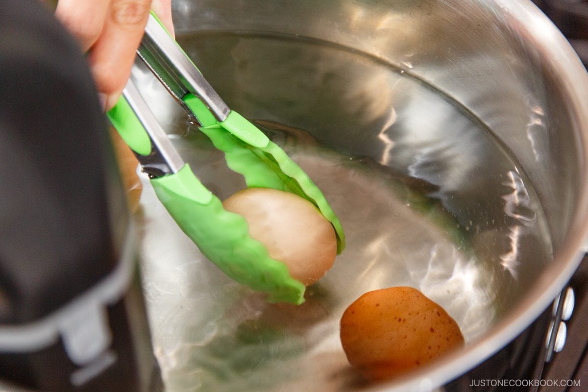 A hand uses green tongs to lift a small potato from a pot of water with other potatoes at the bottom, demonstrating careful handling similar to techniques used in How to Pasteurize Eggs.