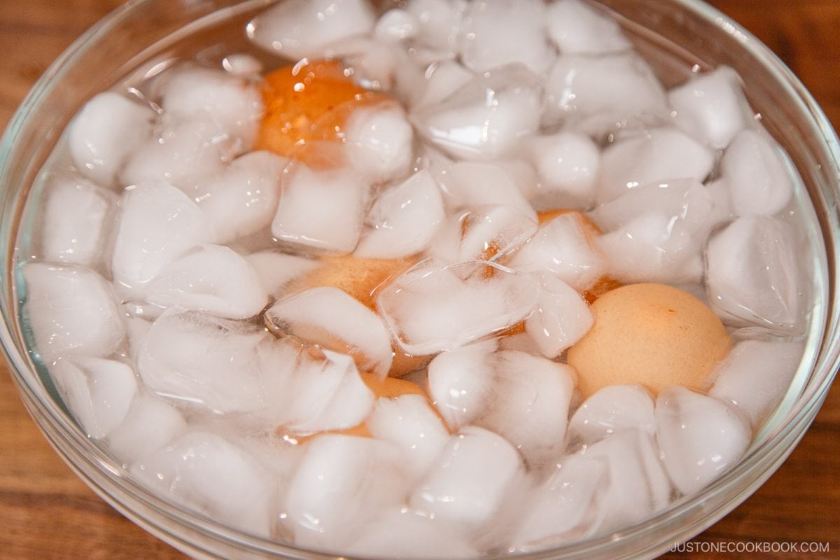 Brown eggs submerged in a clear glass bowl filled with water and ice cubes, creating an ice bath on a wooden surface—a crucial step in the process of how to pasteurize eggs safely at home.