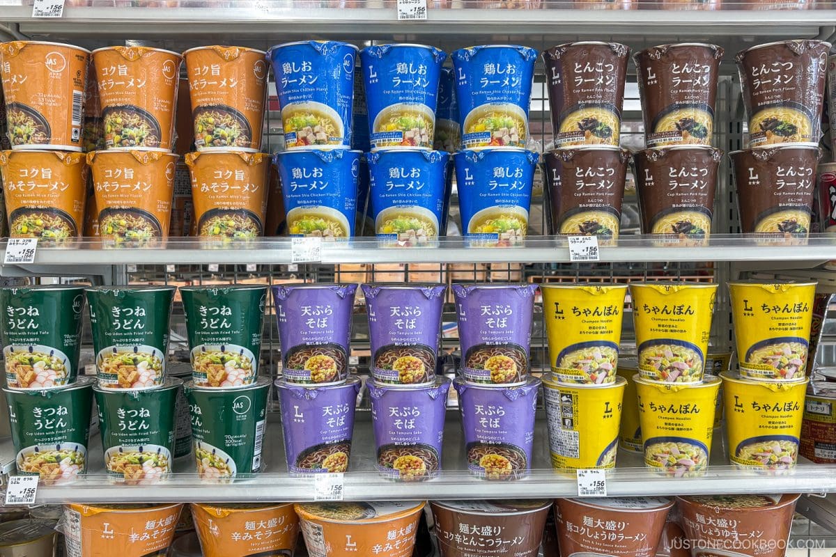 Shelves display colorful instant noodle cups with Japanese labels in orange, blue, brown, green, and yellow, neatly stacked in rows at a grocery store. Price tags are visible under each product&mdash;offering shoppers a tempting selection of the best instant ramen in Japan.