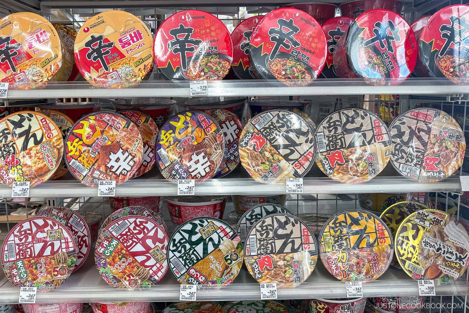Shelves stocked with various colorful instant noodle cups, mostly featuring Japanese and Korean labels, line a grocery store—many showcasing the best instant ramen in Japan. Cups are arranged neatly in rows.