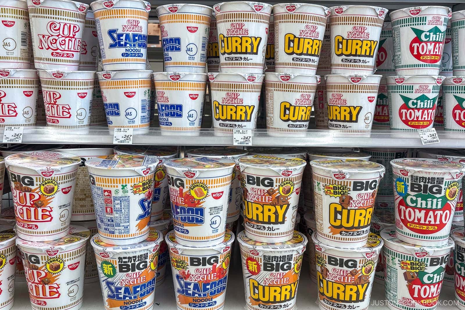 Shelves stocked with various Cup Noodle flavors—including curry, seafood, and chili tomato—in regular and big sizes, showcase some of the best instant ramen in Japan. The colorful packaging is arranged in neat rows, with price tags visible on the shelves below.