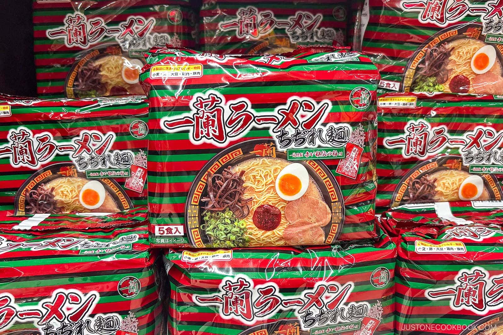 Packets of Ichiran ramen with red, green, and black stripes are stacked together. The packaging shows an image of ramen with pork, egg, and vegetables. Japanese text is printed prominently on the front, highlighting what many consider the best instant ramen in Japan.