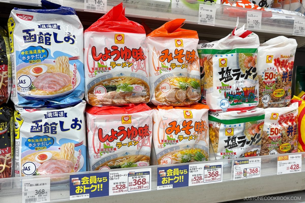 Bags of the best instant ramen in Japan in various colors, with Japanese text and images of noodle bowls, are neatly arranged on a grocery store shelf with price tags displayed below each product.
