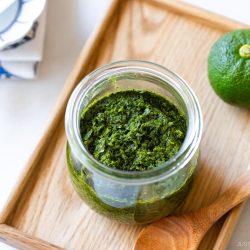 A glass jar filled with vibrant green yuzu kosho paste, made from a traditional Yuzu Kosho Recipe, sits on a wooden tray beside a wooden spoon and a whole green citrus fruit. Plates and utensils are arranged in the background.