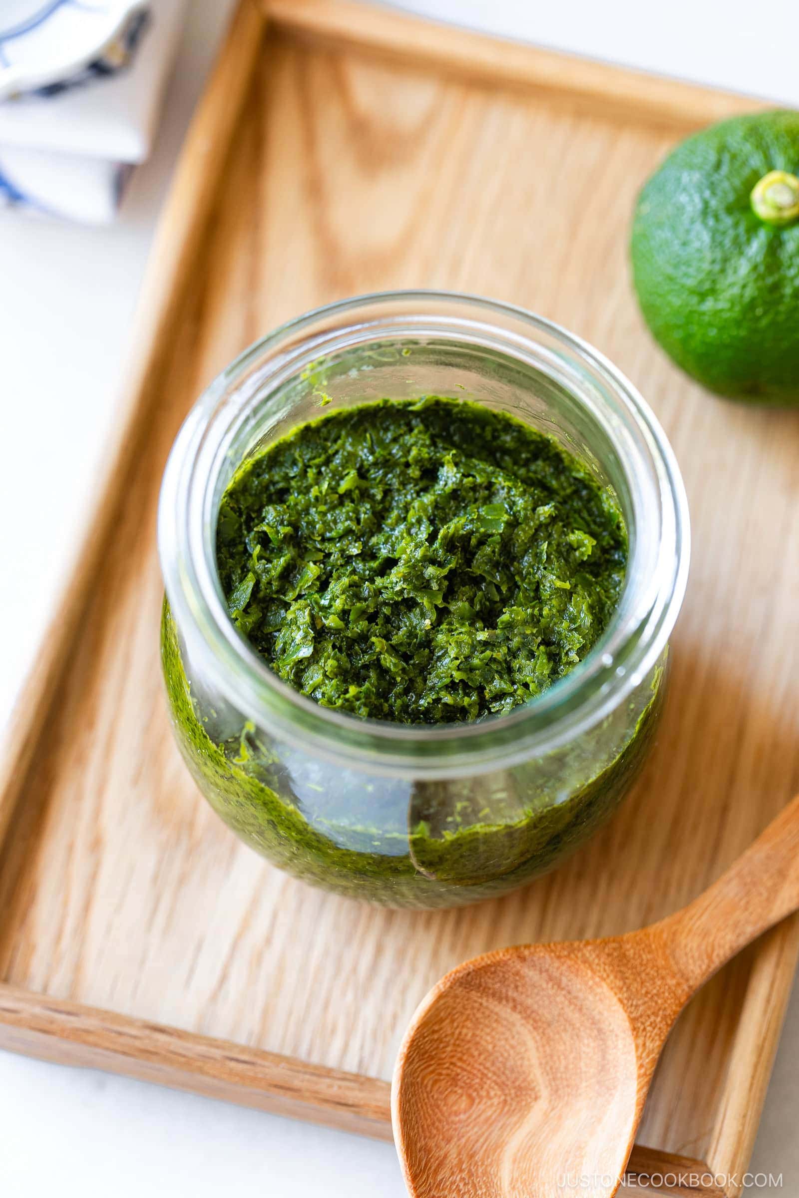 A glass jar filled with vibrant green paste, made using a traditional Yuzu Kosho recipe, sits on a wooden tray next to a wooden spoon and a halved green citrus fruit.