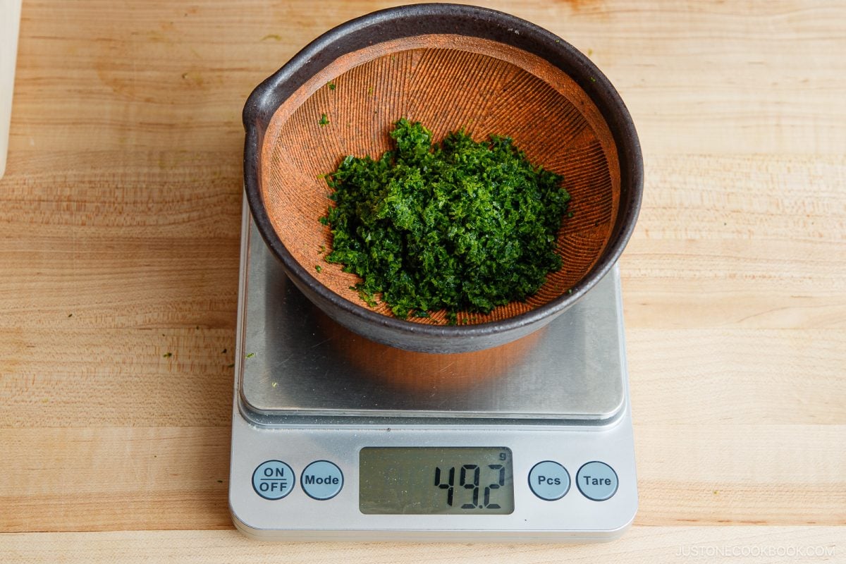 A brown bowl containing finely chopped green herbs, perfect for a Yuzu Kosho recipe, sits on a digital scale reading 49.2 grams, placed on a light wooden countertop.