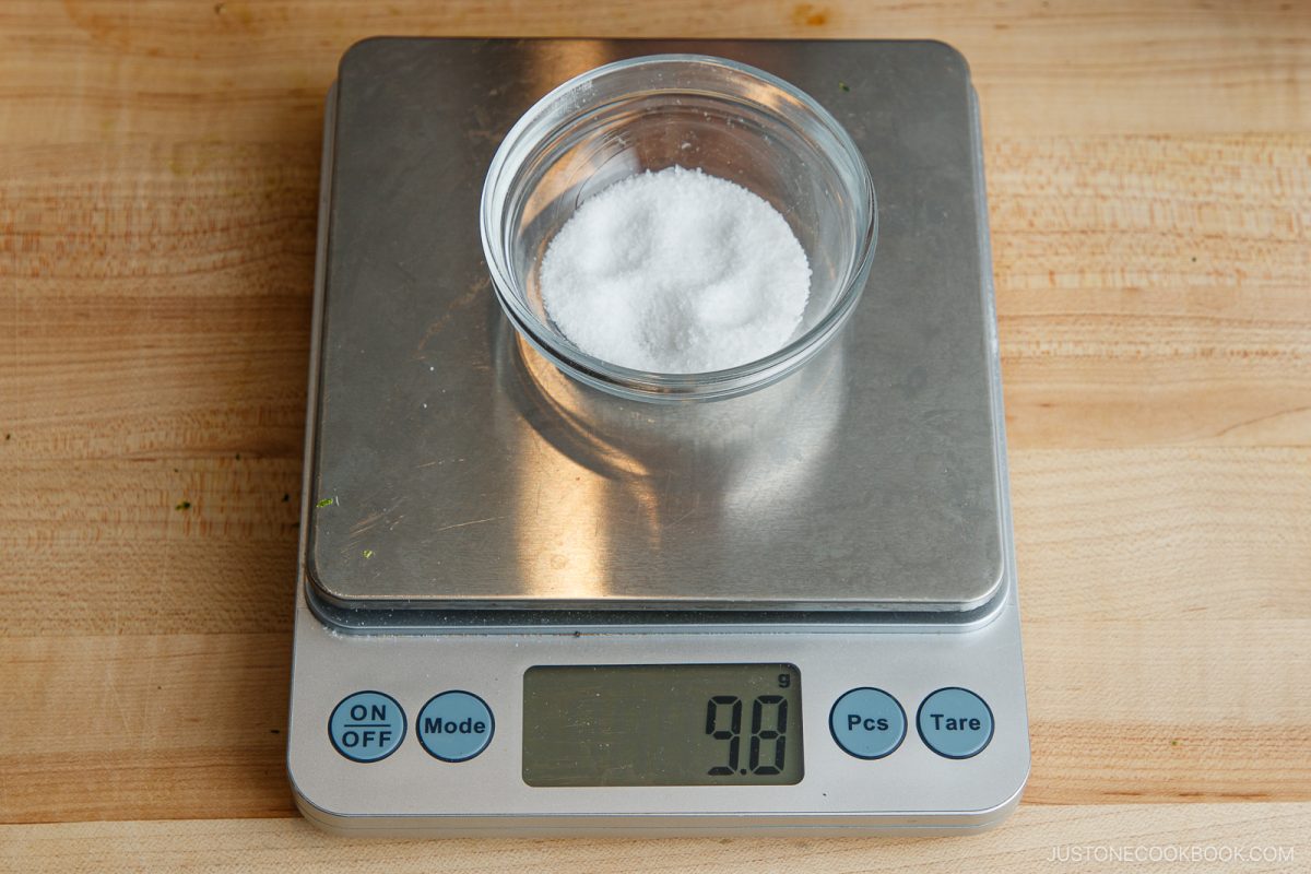 A small glass bowl of white granulated salt sits on a digital kitchen scale, displaying a weight of 9.8 grams, on a wooden countertop&mdash;an essential step in preparing ingredients for your favorite Yuzu Kosho recipe.