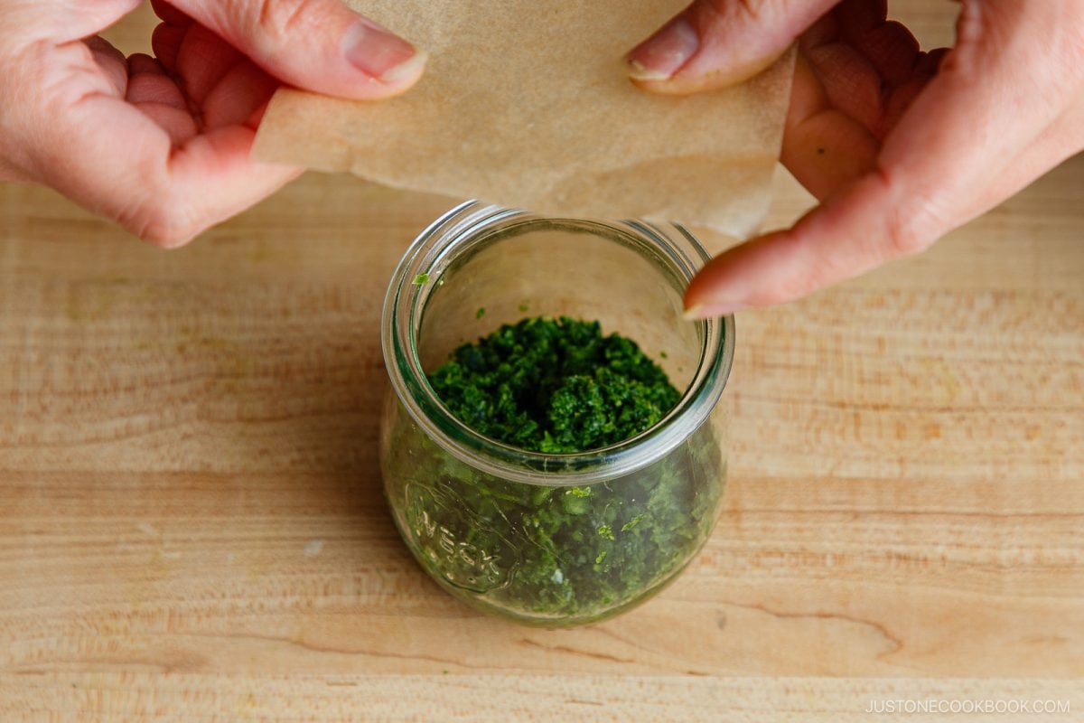 Hands placing a piece of parchment paper on top of chopped green herbs in a glass jar, on a wooden surface&mdash;an essential step in preparing an authentic Yuzu Kosho Recipe.
