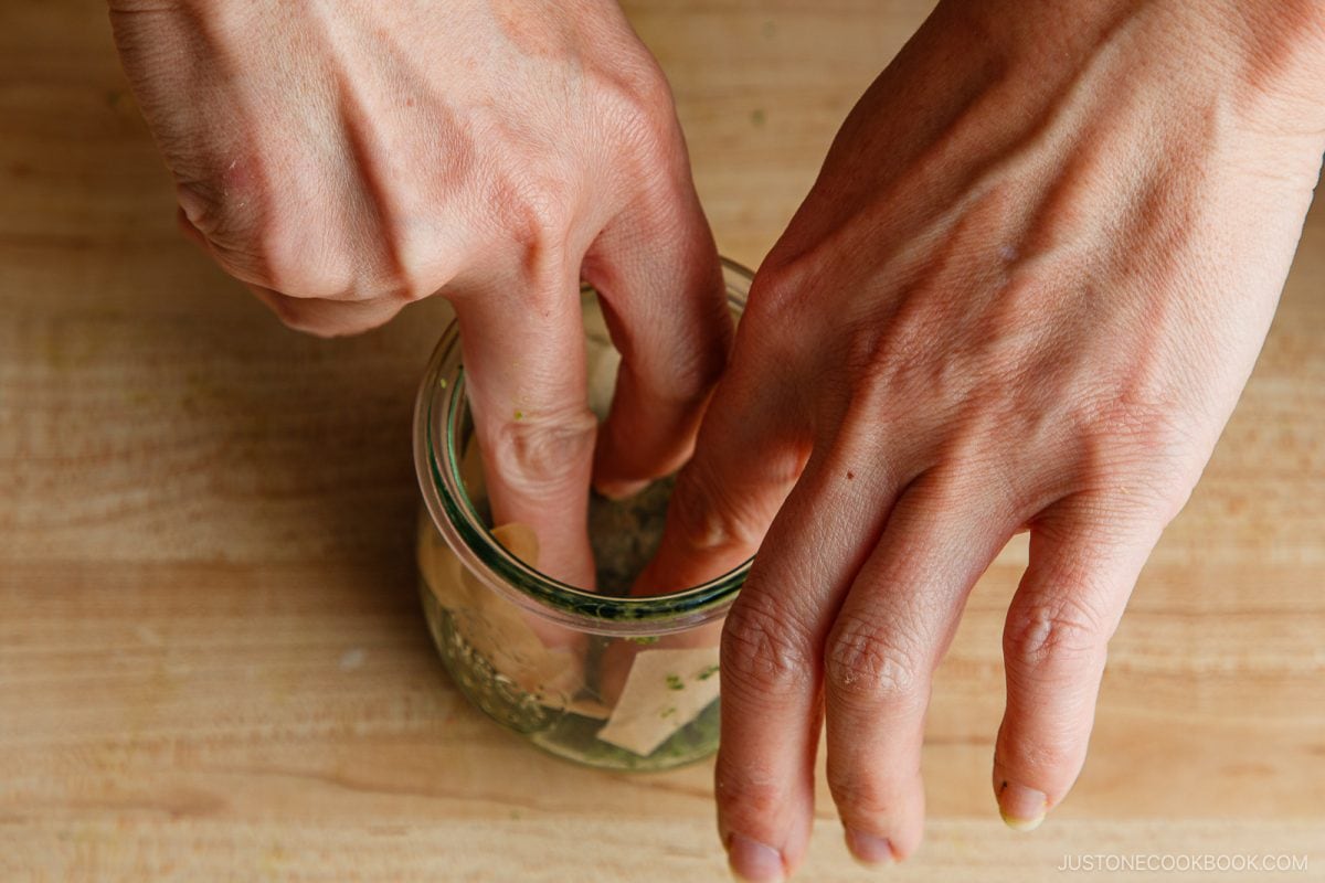 Two hands press cabbage into a glass jar filled with herbs on a wooden surface, likely preparing homemade pickles or kimchi using a Yuzu Kosho recipe.