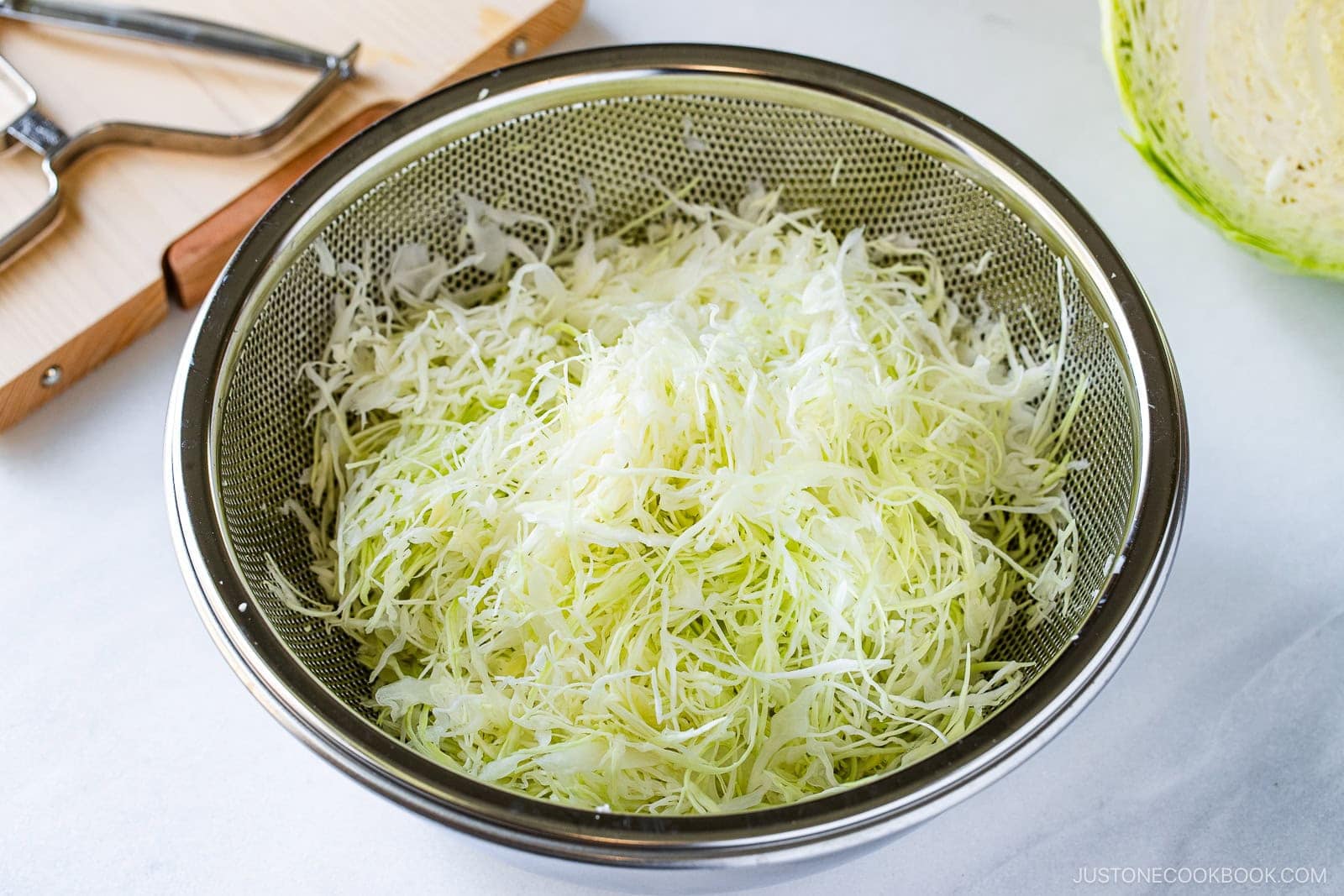 A metal colander filled with finely shredded cabbage, perfect for making a Japanese Shredded Cabbage Salad, sits on a countertop, with a cutting board and kitchen tools in the background.