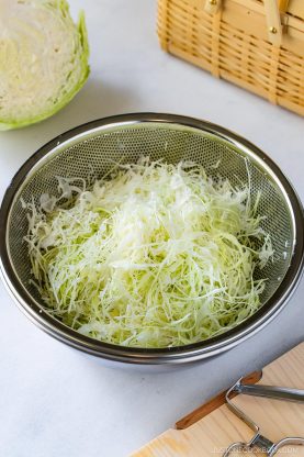 A metal colander filled with thinly shredded cabbage, perfect for Japanese Shredded Cabbage Salad, sits on a white surface, with a cabbage half, a vegetable peeler, and a picnic basket in the background.