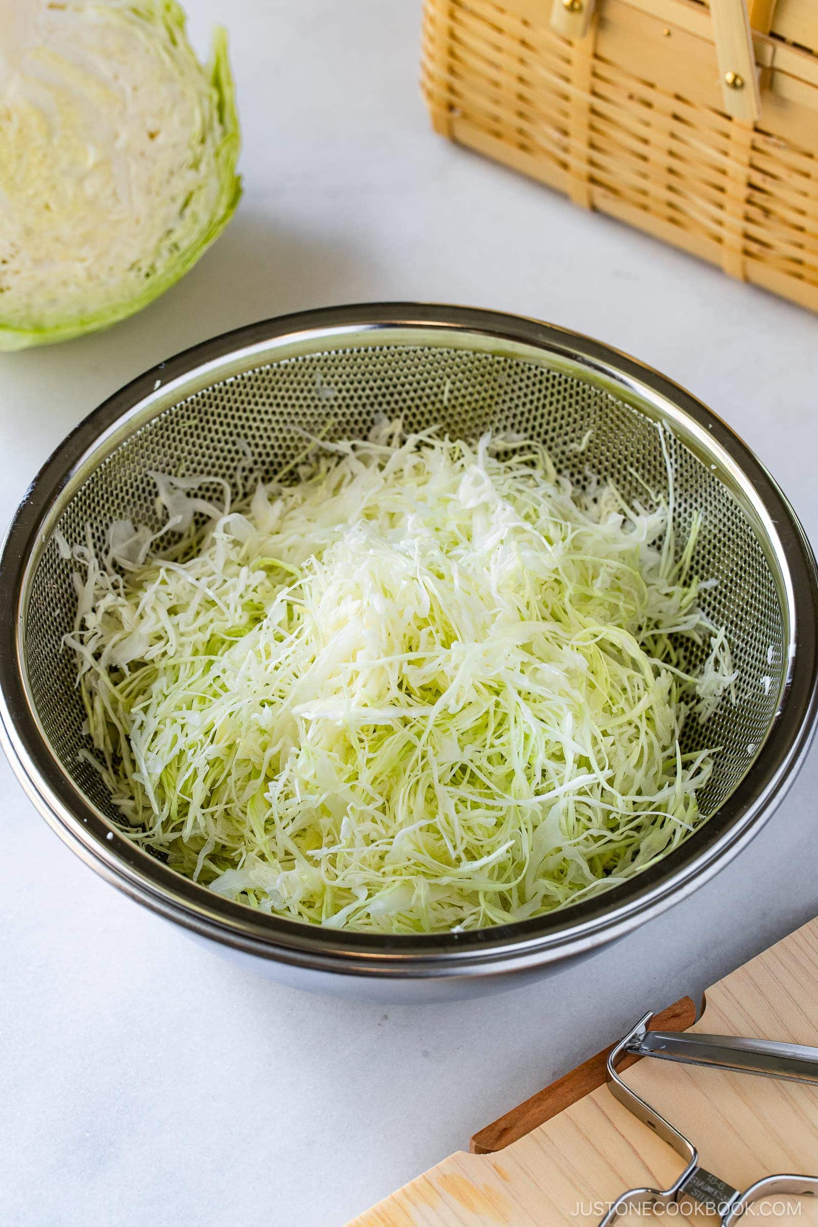 A metal colander filled with thinly shredded cabbage, perfect for Japanese Shredded Cabbage Salad, sits on a white surface, with a cabbage half, a vegetable peeler, and a picnic basket in the background.