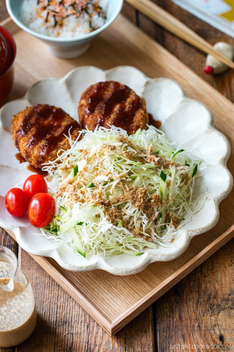 A plate with two breaded cutlets topped with sauce, Japanese Shredded Cabbage Salad with dressing, sliced cucumber, and cherry tomatoes. A bowl of rice and chopsticks are in the background.