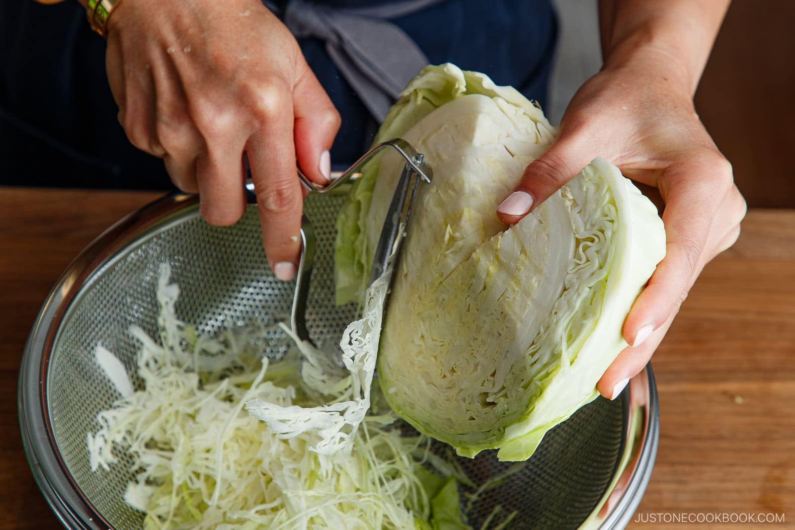 A person prepares Japanese Shredded Cabbage Salad by shredding a head of cabbage over a metal colander using a handheld slicer on a wooden surface.