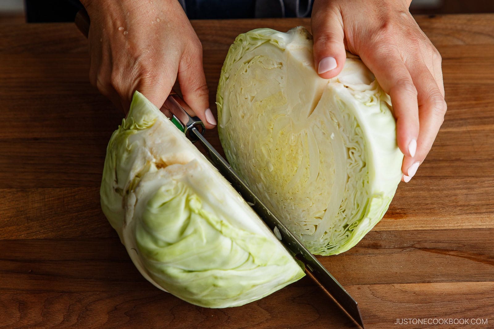 Two hands hold a large knife and cut a green cabbage in half on a wooden surface, preparing fresh ingredients for a Japanese Shredded Cabbage Salad.