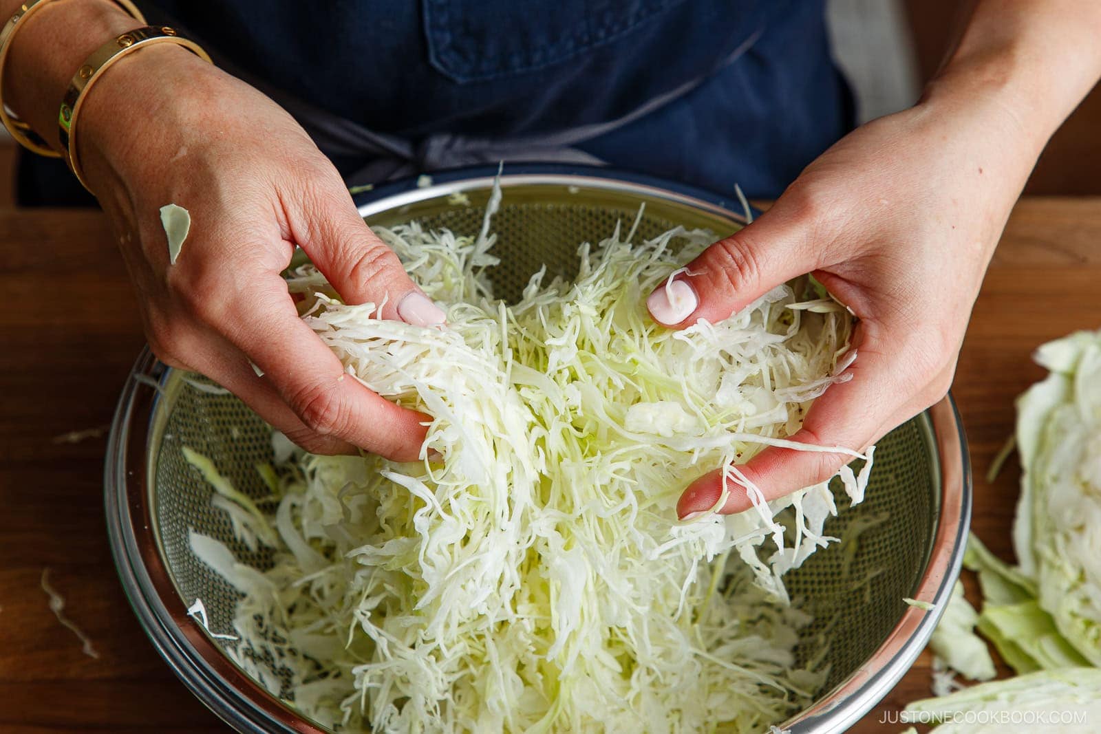Hands holding and mixing freshly shredded cabbage over a metal colander, preparing the base for a Japanese Shredded Cabbage Salad, with a wooden table and a dark apron visible in the background.