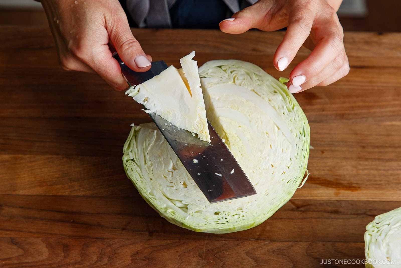 A person slices a wedge from a halved green cabbage on a wooden cutting board using a large knife, preparing fresh ingredients for a Japanese Shredded Cabbage Salad.