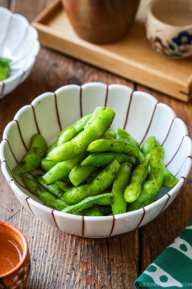 A white bowl with red stripes holds cooked Yuzu Kosho Edamame pods seasoned with black pepper, sitting on a wooden table with small dishes and cups in the background.