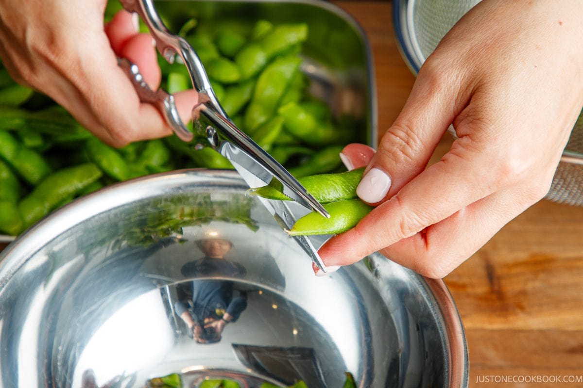 A person uses scissors to trim the ends off a green edamame pod over a stainless steel bowl, preparing fresh Yuzu Kosho Edamame, with more edamame in a tray nearby.