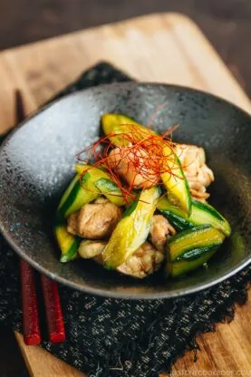 A black bowl filled with stir-fried chicken and sliced zucchini, a delicious Japanese side dish, is garnished with thin red chili threads and sits on a wooden board with red chopsticks and a black textured cloth underneath.