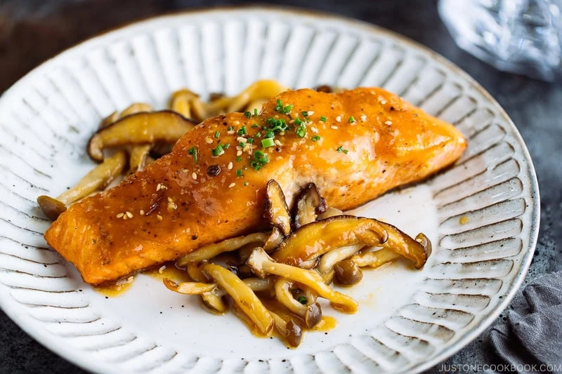 A white plate containing Miso Butter Salmon served with saut&eacute;ed shiitake and shimeji mushrooms.