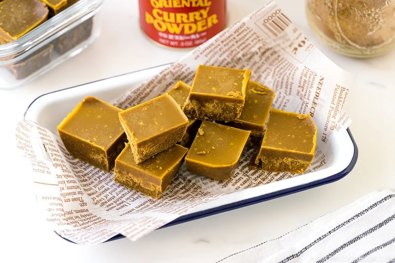 A white tray lined with newspaper-style paper holds several square pieces of curry-flavored fudge reminiscent of Japanese chicken curry. A jar labeled curry powder and a glass container with more fudge are in the background.