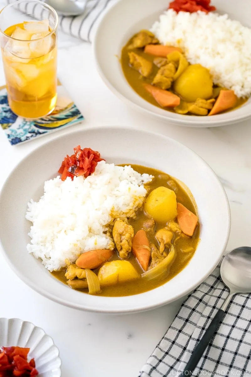 A bowl of Japanese chicken curry with tender pieces of chicken, potatoes, carrots, and white rice, garnished with red pickles. An iced drink, a spoon, and a striped napkin are set beside the bowl. Another bowl is partially visible in the background.