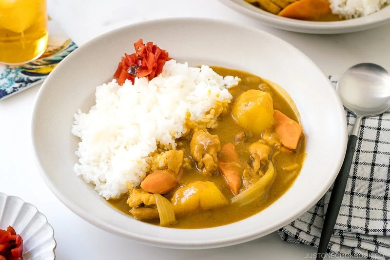 A plate of Japanese chicken curry with rice, featuring tender chicken, potatoes, carrots, onions, and a garnish of red pickles, served with a spoon on a checkered napkin.