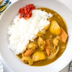 A plate of Japanese chicken curry with white rice, topped with red pickles. The curry contains chunks of chicken, carrots, and potatoes. A spoon and napkin are placed to the right of the plate.