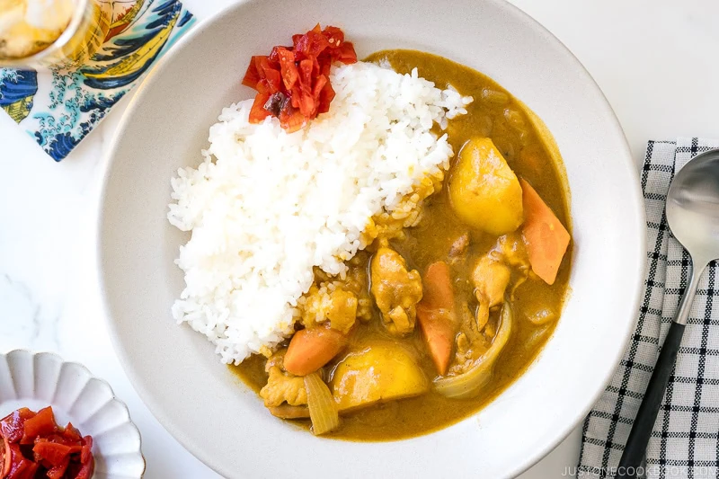 A plate of Japanese chicken curry with tender pieces of chicken, potatoes, carrots, and onions is served with white rice and a small portion of red pickles on top. A spoon and napkin are placed beside the dish.