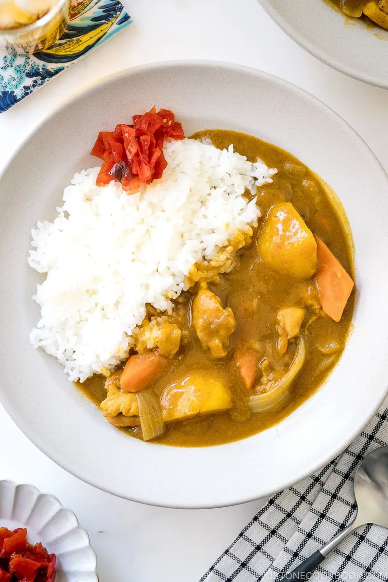A white plate with Japanese chicken curry, steamed white rice, and red pickled ginger. The curry contains chunks of chicken, carrots, and potatoes. A spoon and a small bowl of extra ginger are beside the plate.