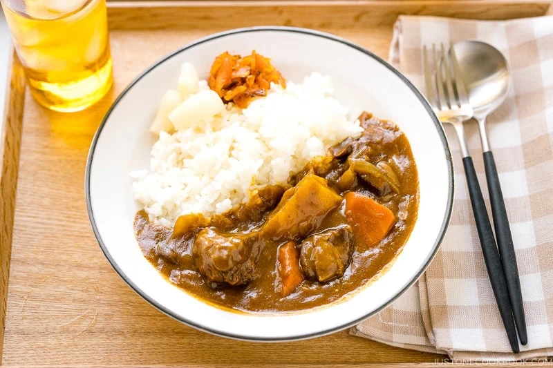 A plate of Japanese chicken curry with rice, carrots, potatoes, and tender pieces of chicken is served alongside pickled vegetables. A glass of iced tea and a spoon and fork on a napkin are placed beside the plate on a wooden tray.