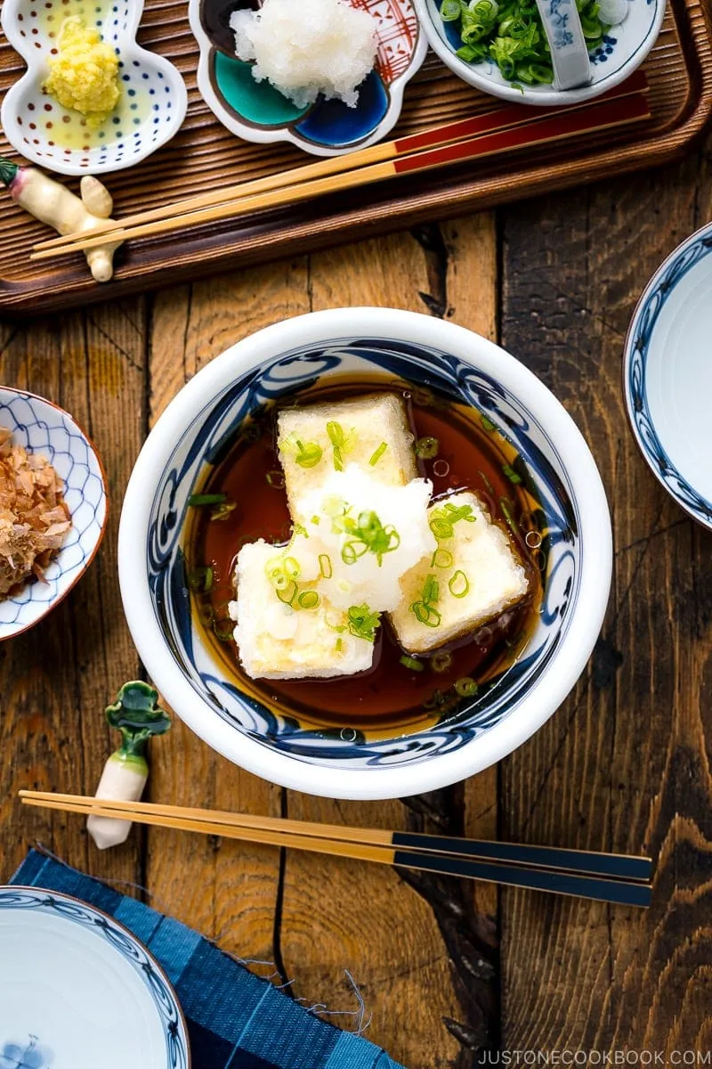 A ceramic bowl containing Agedashi Tofu (Agedashi Dofu) topped with grated daikon, chopped scallions, and grated ginger.