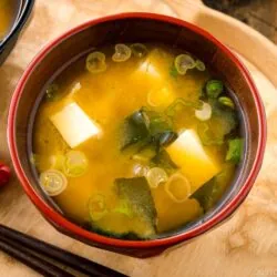 A bowl of miso soup with tofu cubes, seaweed, and chopped green onions sits on a wooden tray next to chopsticks and a red flower-shaped dish&mdash;a classic presentation for any authentic miso soup recipe.