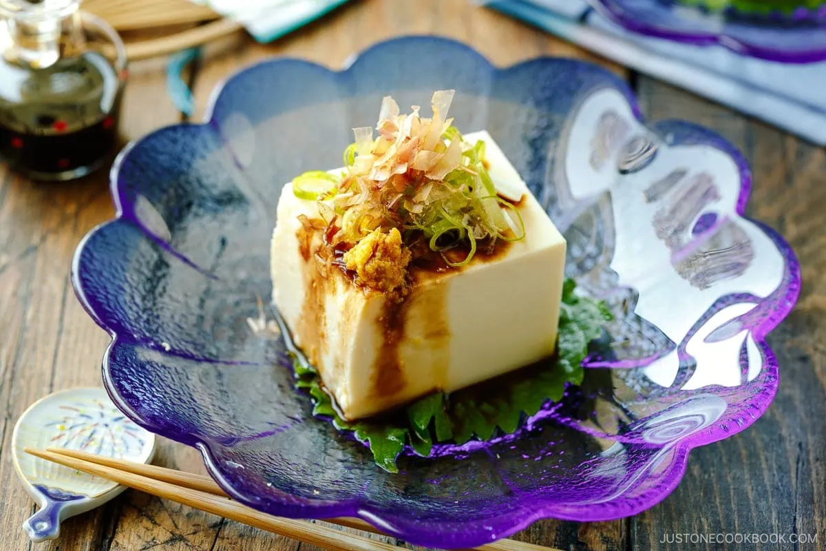 A glassware containing Japanese cold tofu topped with green onion, grated ginger, and katsuobushi and drizzled with soy sauce.