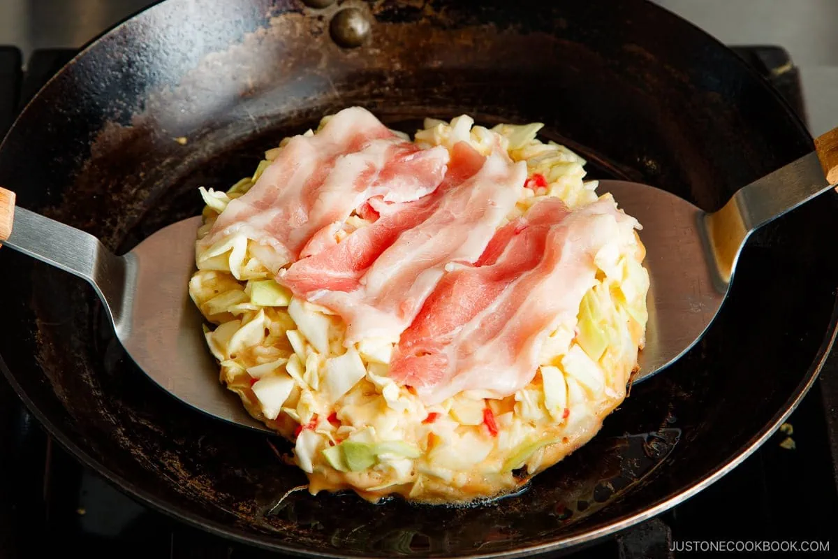 A round okonomiyaki cabbage pancake topped with raw pork slices is being cooked in a black skillet, held by two wide metal spatulas on either side. The okonomiyaki pancake is golden at the edges.