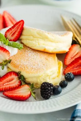 A white plate containing Japanese souffle pancakes dusted with powdered sugar, fresh mixed berries, and whipped cream.