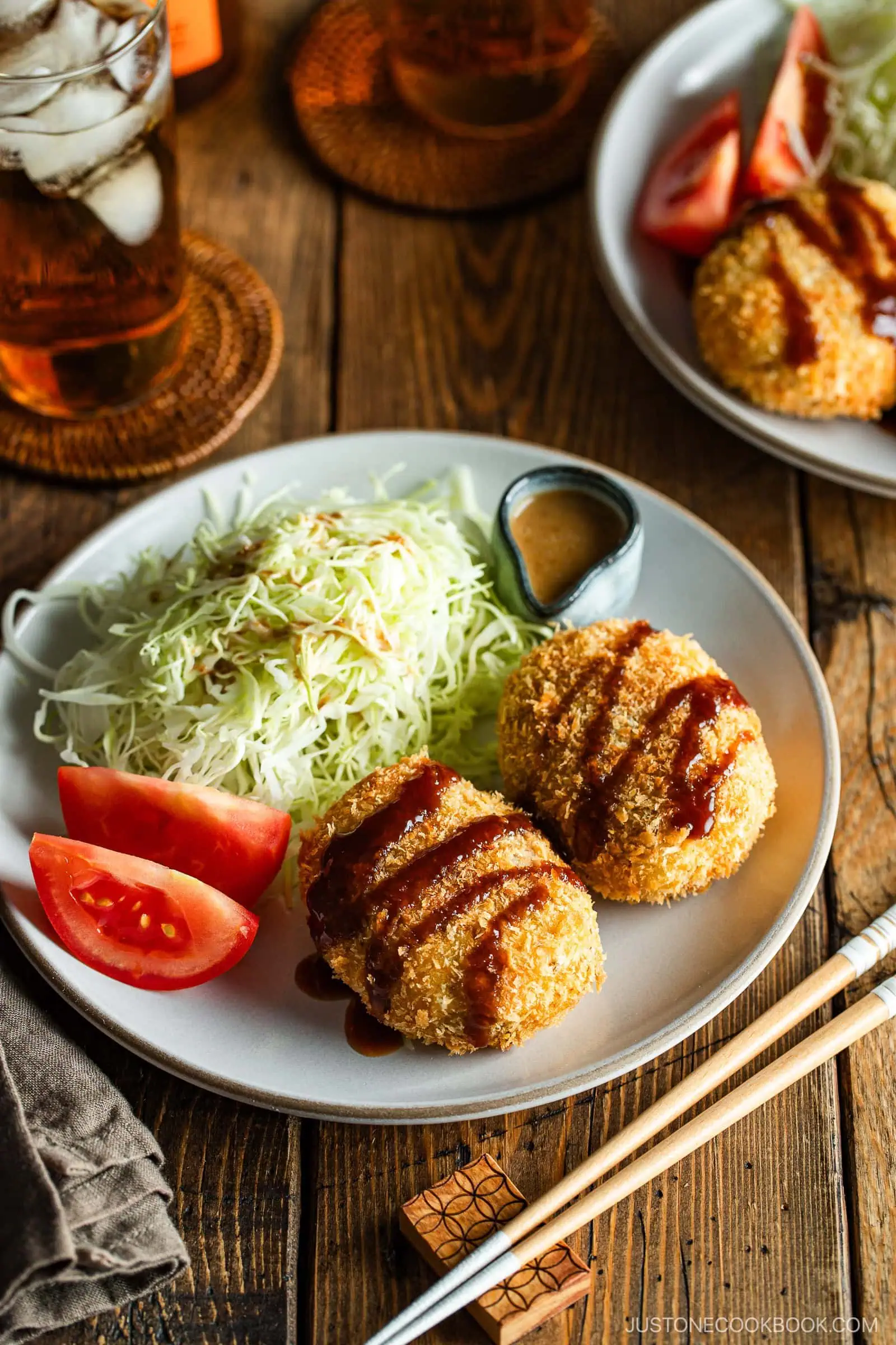 A round white plate containing Japanese croquettes, shredded cabbage, tomato wedges, and a tiny container with sesame dressing.