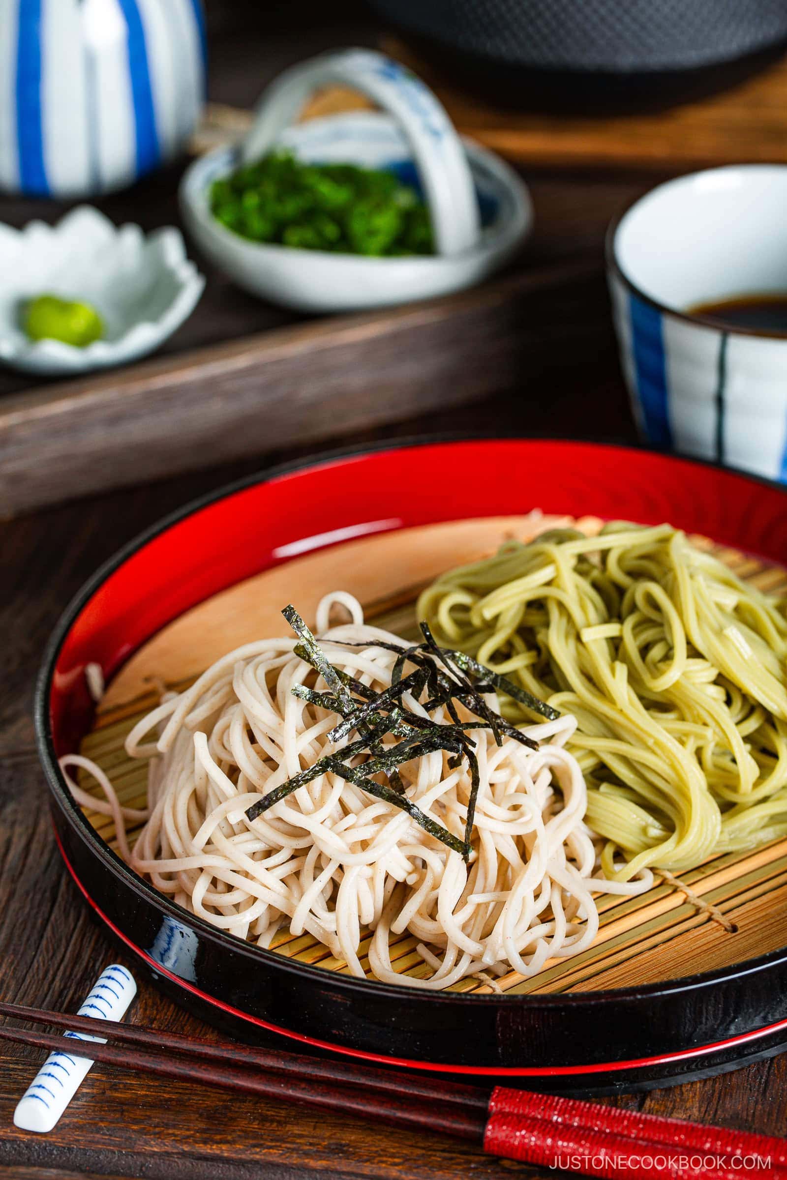 A red and black tray presents Zaru Soba (Cold Soba) in two bundles—one light, one green—garnished with shredded nori, served on a bamboo mat, with dipping sauce, chopped green onions, and wasabi in the background.