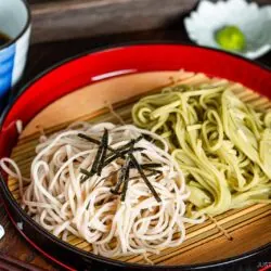 A red tray of Zaru Soba (Cold Soba) features white and green soba noodles topped with sliced seaweed, placed on a bamboo mat beside a bowl of dipping sauce, wasabi, and chopped scallions. Chopsticks rest alongside the tray.