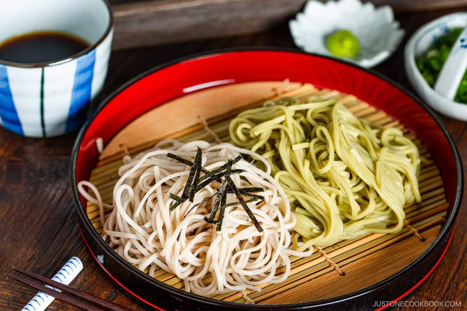 A red tray of Zaru Soba (Cold Soba) features white and green soba noodles topped with sliced seaweed, placed on a bamboo mat beside a bowl of dipping sauce, wasabi, and chopped scallions. Chopsticks rest alongside the tray.