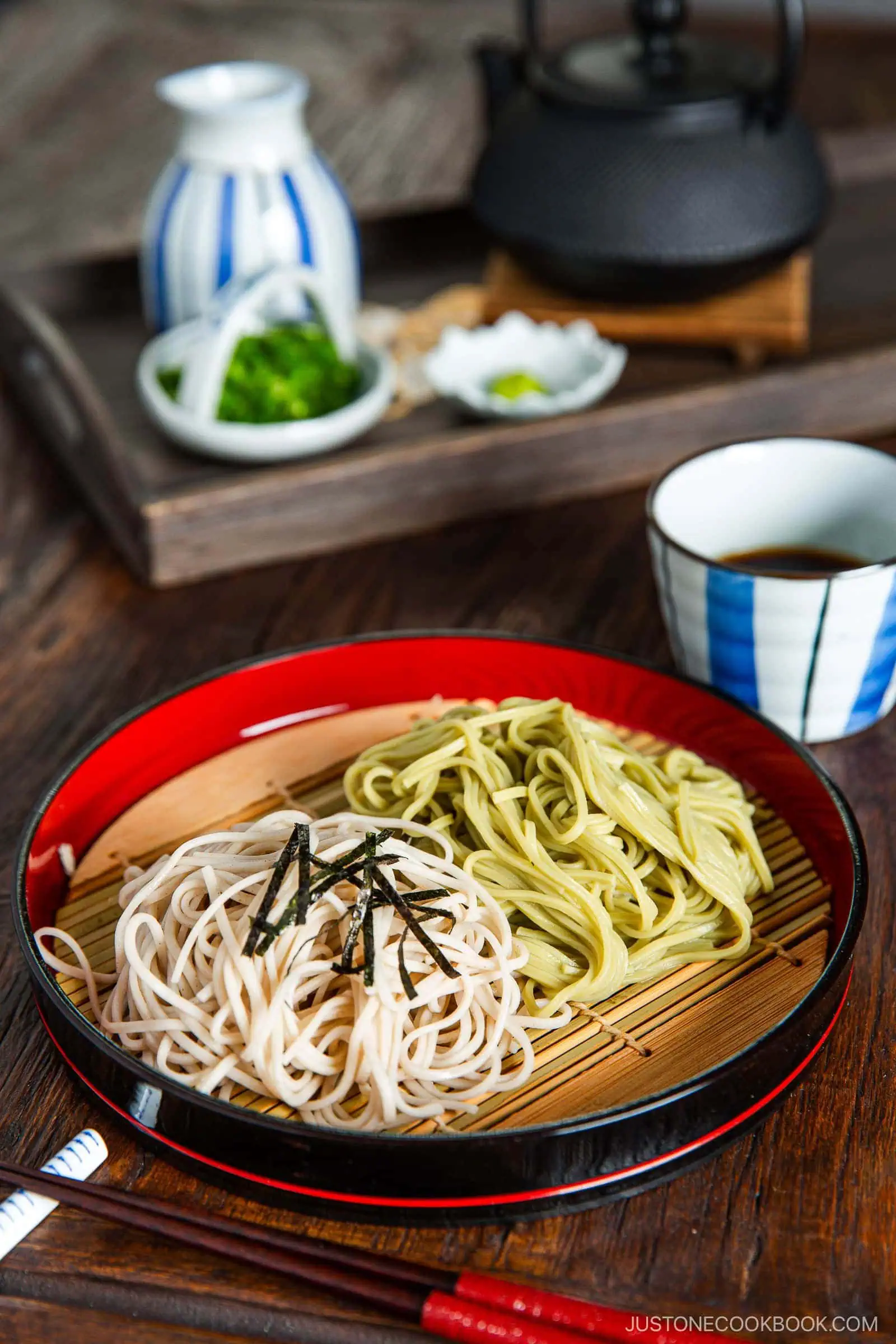 A plate with two types of Japanese Zaru Soba (Cold Soba) noodles—regular zaru soba and green tea soba, garnished with shredded seaweed. A dipping sauce, teapot, and condiments are in the background on a wooden tray.