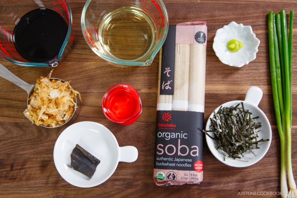 A variety of ingredients for making Zaru Soba (Cold Soba) are arranged on a wooden surface, including dried soba noodles, soy sauce, dashi, mirin, bonito flakes, kombu, sliced green onions, nori strips, and wasabi.