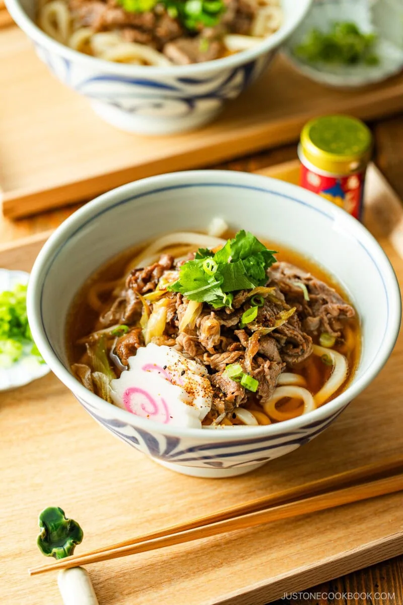 A bowl of udon noodle soup topped with sliced beef, fish cake, green onions, and cilantro, set on a wooden table with chopsticks and a condiment container nearby.
