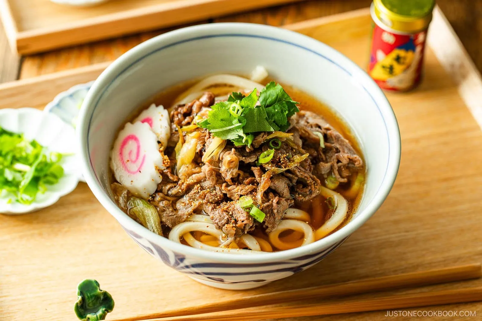 A bowl of beef udon with sliced beef, green onions, narutomaki (fish cake), and fresh herbs in broth, served on a wooden tray with chopped green onions and a bottle of seasoning on the side.