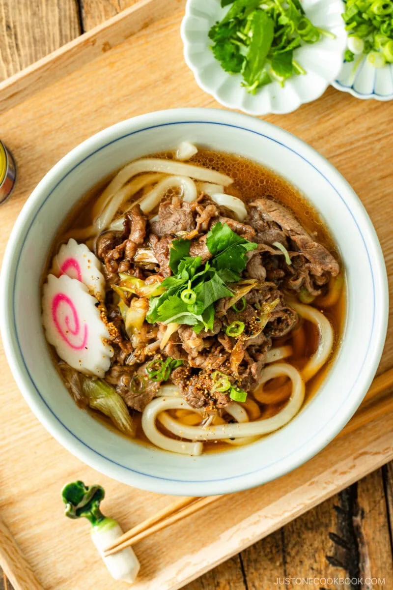 A bowl of udon noodle soup topped with sliced beef, narutomaki fish cake, green onions, and fresh herbs, served on a wooden tray with extra green onions and a pair of chopsticks on the side.